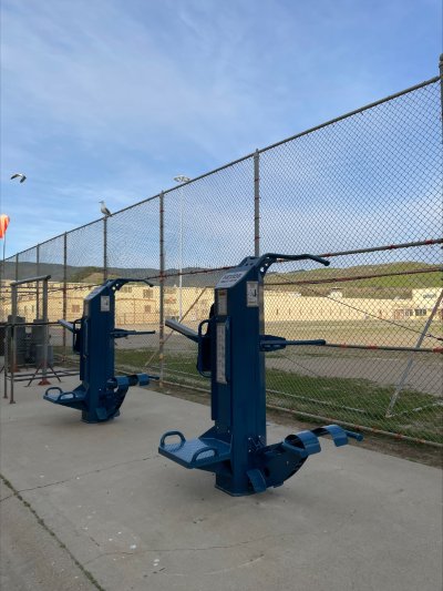 Correctional fitness equipment installed in an outdoor prison recreation yard with inmates participating in structured exercise.